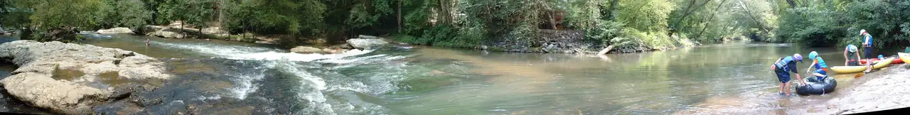 An ultra-wide summer view of a shallow river: pale granite ledges and a white-water rapid on the left, a calm green-brown pool in the middle under a wall of leafy trees, and on the right a sandy rock bank where two children in helmets and life jackets ready a black inner tube beside yellow and red kayaks and two adults.