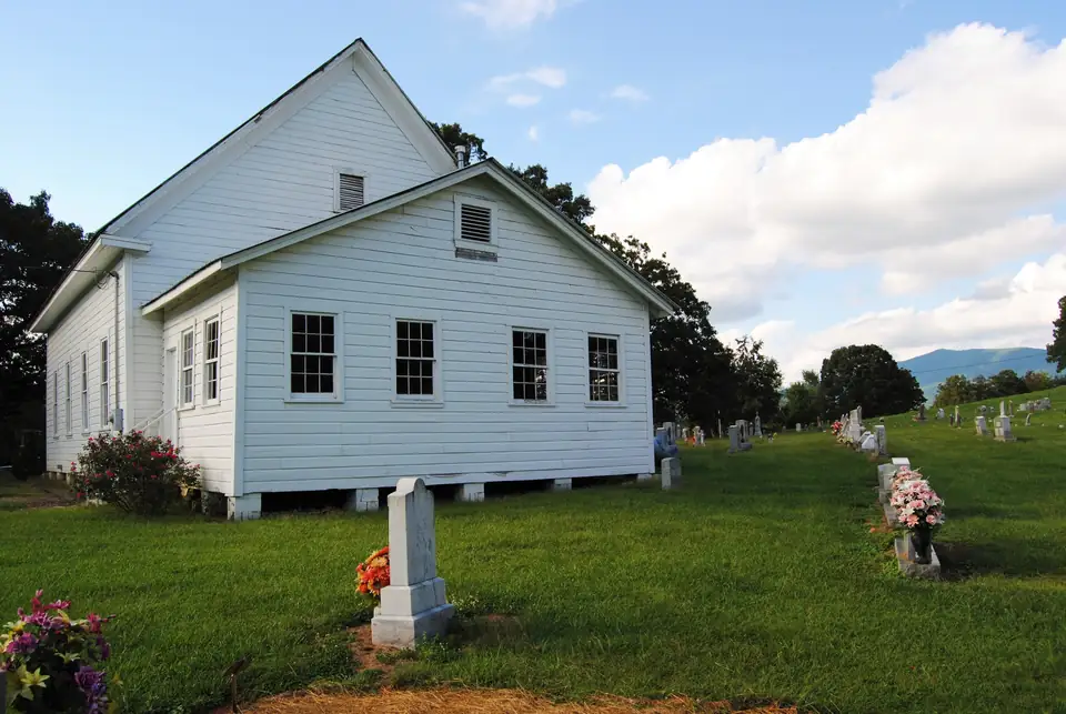 A white clapboard church with a front gable and a lower rear wing stands on a green lawn among rows of gravestones decorated with artificial flowers, under a blue sky with large white clouds, with a blue mountain ridge on the right horizon.