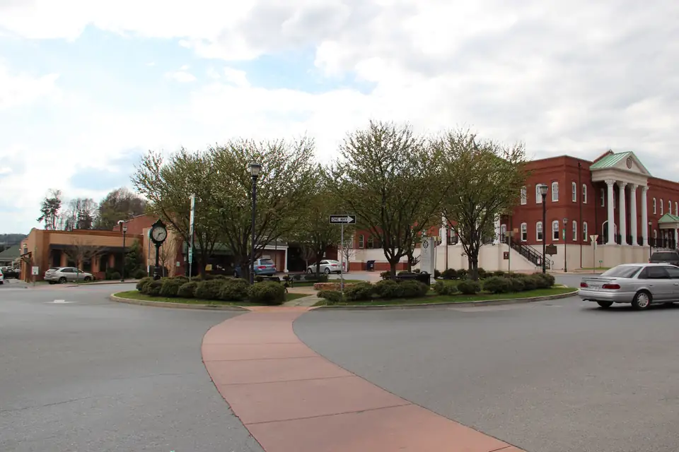 A curving brick-edged road loops around a landscaped roundabout with young trees and a clock post, with the white-columned red-brick courthouse and storefronts behind and a silver car on the road, under a cloudy sky.