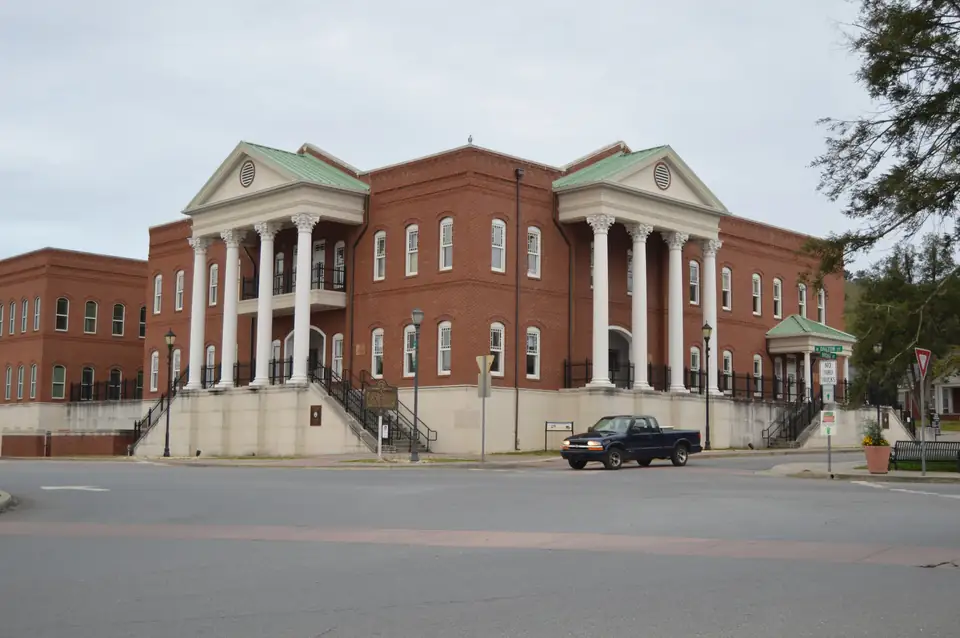 A large red-brick courthouse with two white-columned porticoes and green-roofed pediments stands across an empty street under an overcast sky, with a dark pickup truck parked in front.