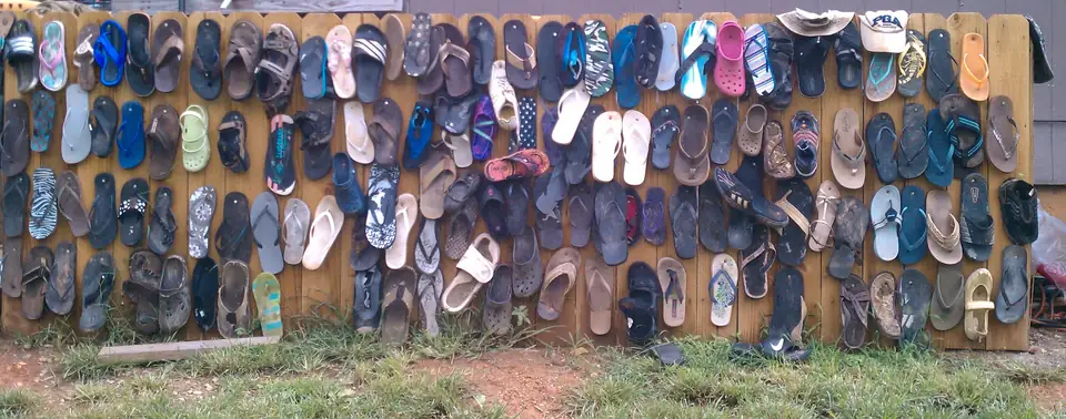 Rows of mismatched flip-flops, sandals, water shoes and two caps are hung on a wooden board fence in front of a grey wooden building, above a strip of patchy grass and red clay.