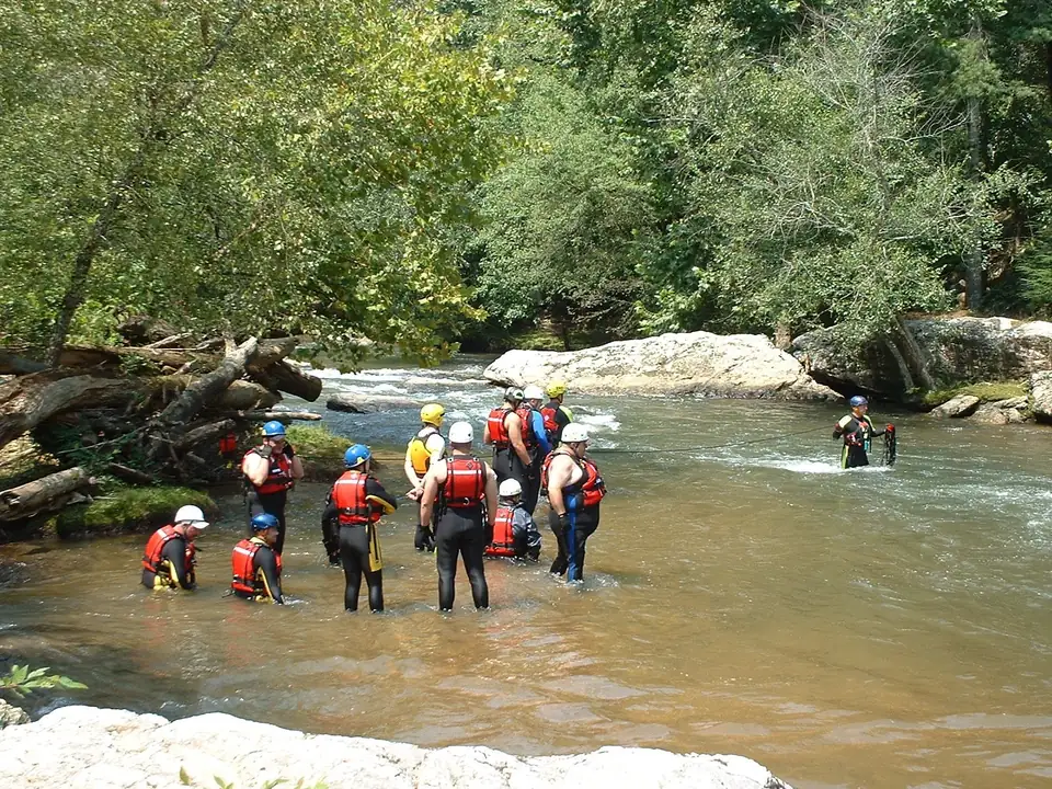 About a dozen people in wetsuits, red life jackets and white, blue and yellow helmets stand knee- to waist-deep in a brown river below a small rapid, with a rope stretched across the current to a rescuer by a large pale boulder, fallen logs on the left bank and leafy trees behind.