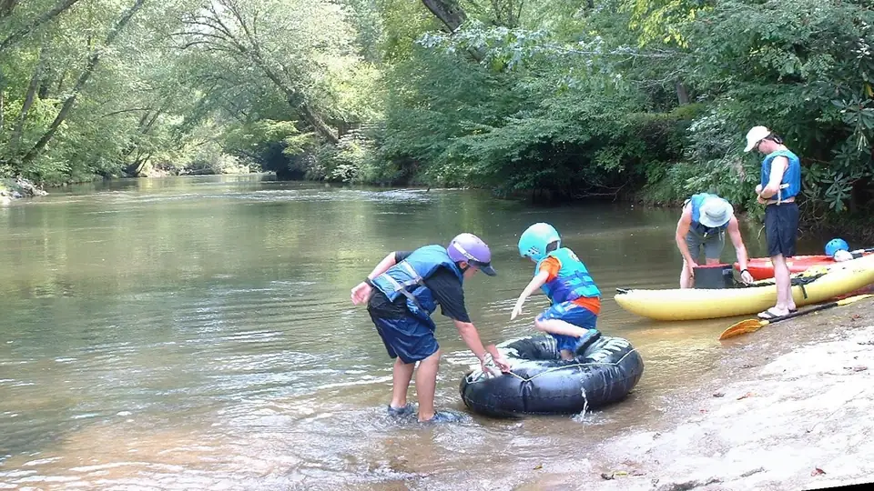 Two children in helmets and life jackets climb onto a black inner tube in shallow brown water at a sloping rock bank, while two adults in life jackets and sun hats stand by yellow and red kayaks, with the calm river curving away under arching summer trees.