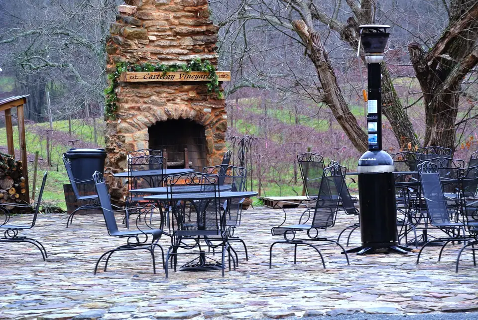 A flagstone patio with black wrought-iron tables and chairs, a tall patio heater and a big stone outdoor fireplace hung with a garland and a wooden sign reading "est. Cartecay Vineyards 2007", with bare trees and rows of leafless vines on a green slope behind.